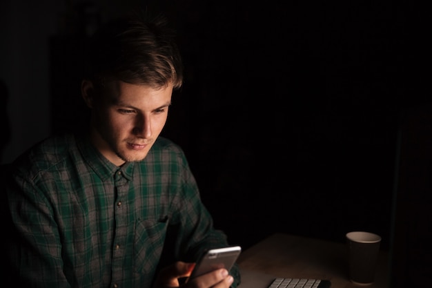 Serious young man sitting and using cell phone in dark room