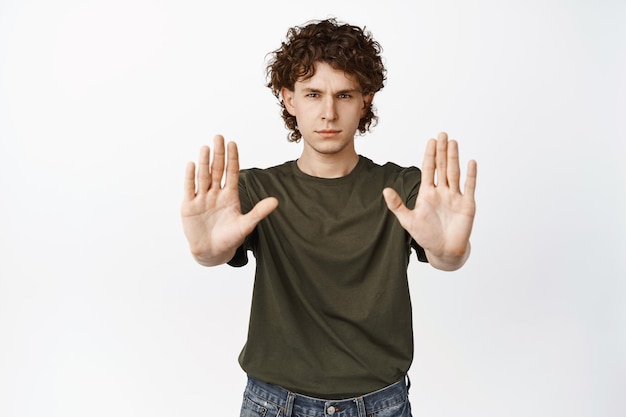 Serious young man stretch out hands showing stop prohibition gesture forbid smth bad standing over white background