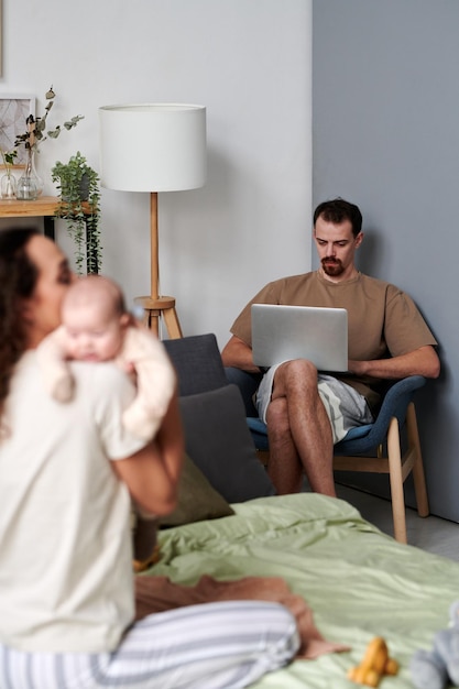 Serious young man with laptop networking in front of his wife with baby son