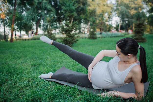 Serious young pregnant woman stretching legs on yoga mate in park. She hold one hand on belly. Right legs is in air.