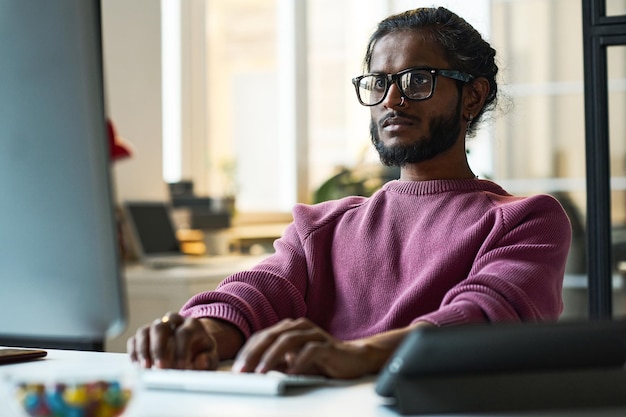 Serious young programmer in eyeglasses working online on computer at his workplace in it office