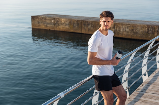 Serious young sportsman standing and drinking water on pier