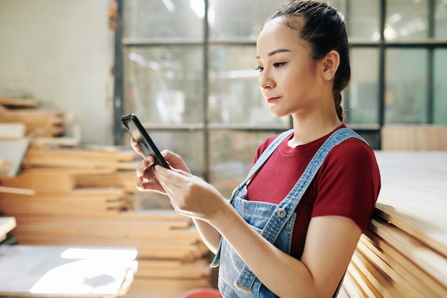 Serious young Vietnamese female carpenter checking text messages in her smartphone from customers or friend