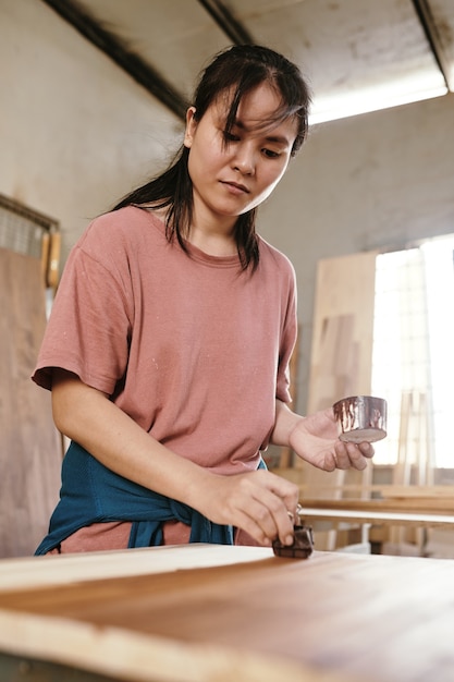 Serious young woman applying lacquer to protect and give shine to sanded wooden board