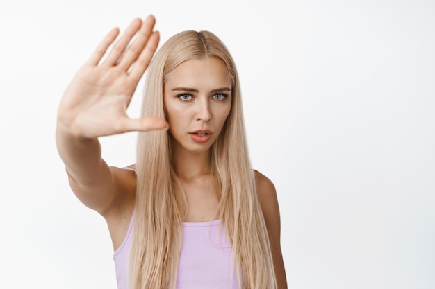 Serious young woman asking to stop filming her, showing no, rejection gesture, refusing and say no, standing on white