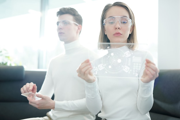 Photo serious young woman in smart goggles analyzing scheme on glassy device while her boyfriend using transparent control panel