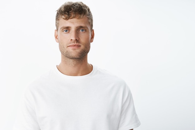 Seriouslooking peaceful and calm attractive blond european man with blue eyes and bristle looking at camera focused and chill without emotions unbothered over gray background in white tshirt