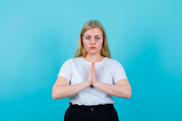 Seriously blonde girl is looking at camera by holding handfuls together on chest on blue background