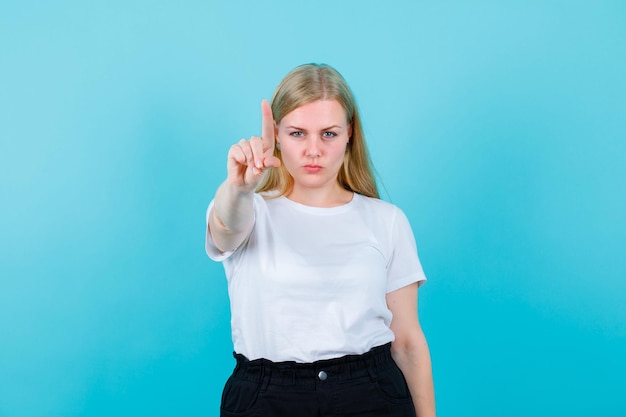 Seriously blonde girl is showing aminute gesture on blue background