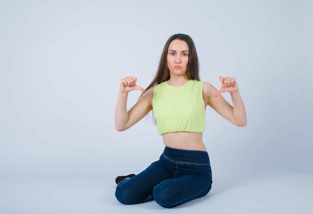 Seriously girl is pointing down with thumbs by sitting on floor on white background