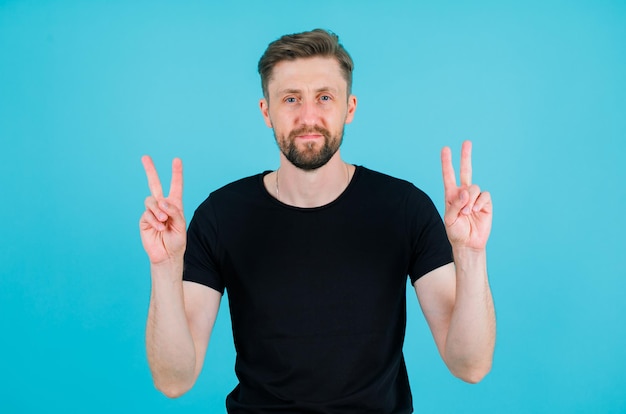 Seriously young man is looking at camera by showing two gestures on blue background