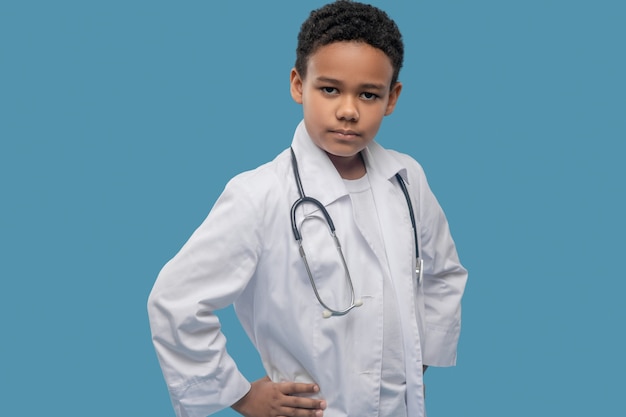 Seriousness. Serious involved dark skinned boy in white medical coat and stethoscope posing self mportant