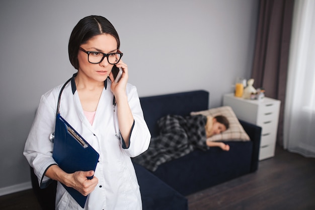 Serius worried young woman talk on phone. She stand in room. Her patient lying on couch behind. Small sick girl sleep.