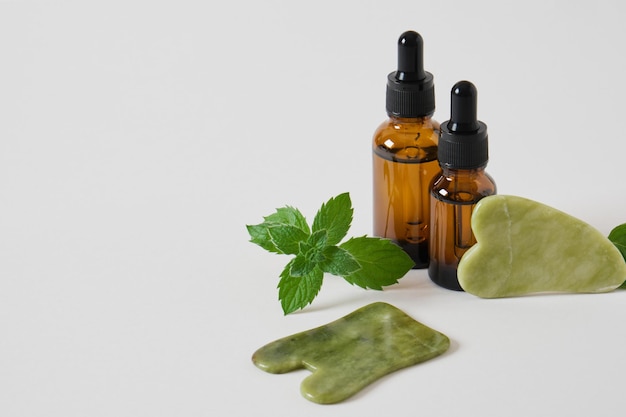 Serum or cosmetic oil in amber glass dropper bottles and a gua sha scrapers made of jadeite stone and mint leaves on gray background