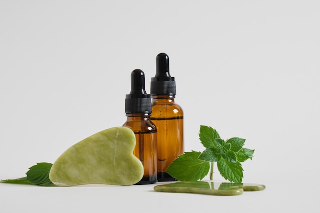 Serum or cosmetic oil in amber glass dropper bottles and a gua sha scrapers made of jadeite stone and mint leaves