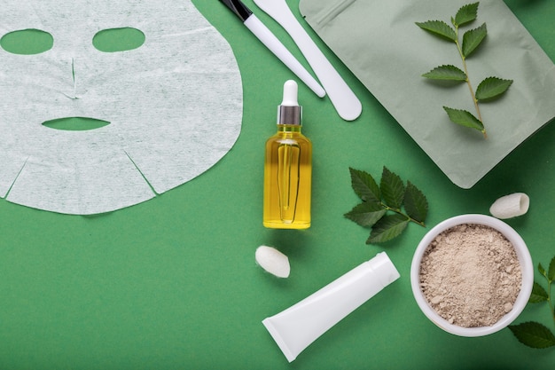 Serum oil and fabric cosmetic face mask with set cosmetics clay mask spatula brush, moisturizing cream. Beauty spa treatments for facial skincare, cosmetology on green background with leaves.