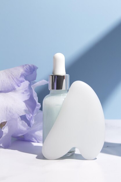 Serum with floral extracts for skincare Gua sha stone for face massage Nature cosmetics in glass bottle with pipette and iris flowers on blue background Face and body care spa concept