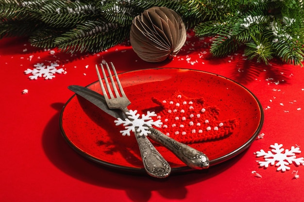 Served Christmas table setting in red tones with vintage cutlery. Evergreen branches, crocheted fir trees, artificial snow. Modern hard light, dark shadow. Flat lay, red background, close up