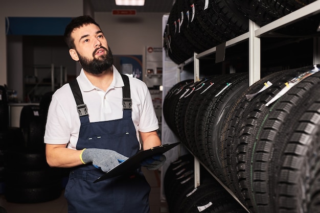 Service staff bearded man examining tires in a supermarket mall, think about right trade for the size of the wheel, holding paper tablet in hands, checking