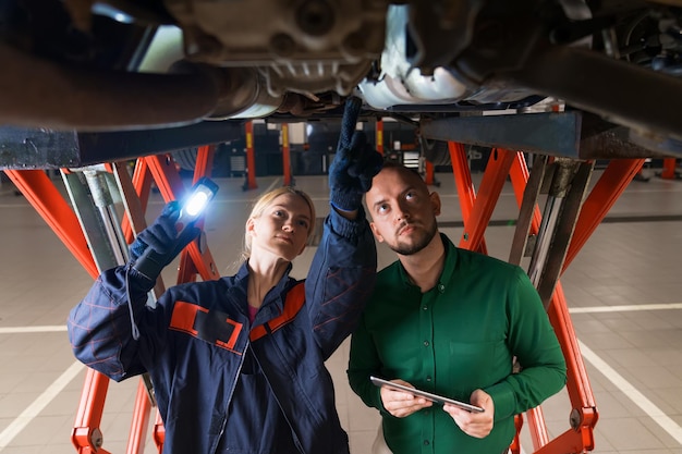 The service station manager writes down in the tablet what needs to be repaired in the car A young female mechanic with a flashlight is standing nearby Car on the lift