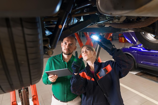 A service station manager and a young female car mechanic are standing under a car on a lift Diagnostics of the suspension and steering of the car