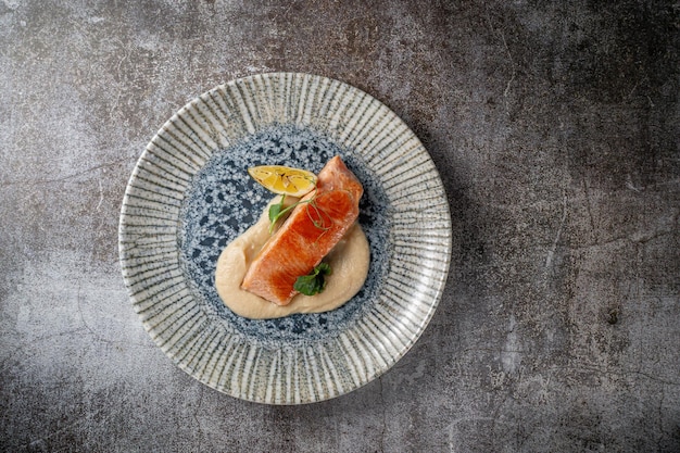 Serving a dish from a restaurant menu. Baked salmon steak with lemon, salad and dill and parsley greens on a plate against a gray stone table