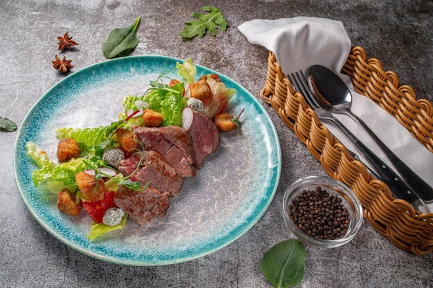 Serving a dish from the restaurant menu. Meat fried al dente (Semi-cooked) with vegetables, green salad and croutons on a plate against the background of a gray stone table, a delicious appetizer