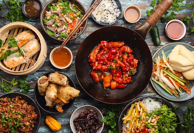 Set of assorted Chinese dishes on table: sweet and sour chicken in wok pan, dim sum in bamboo steamer, spring rolls, noodles, salad, rice, steamed buns, dips. Asian style dinner or buffet, top view