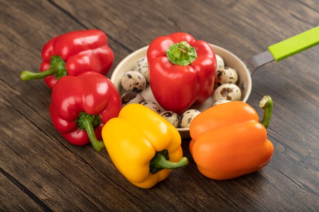 Set of colorful sweet bell peppers and pan of quail eggs on wooden surface