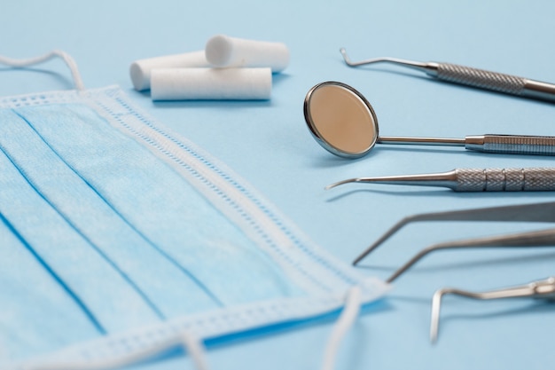 Set of composite filling instruments for dental treatment and protective mask. Medical tools on blue background with copy space. Focus on a mirror. Shallow depth of field.