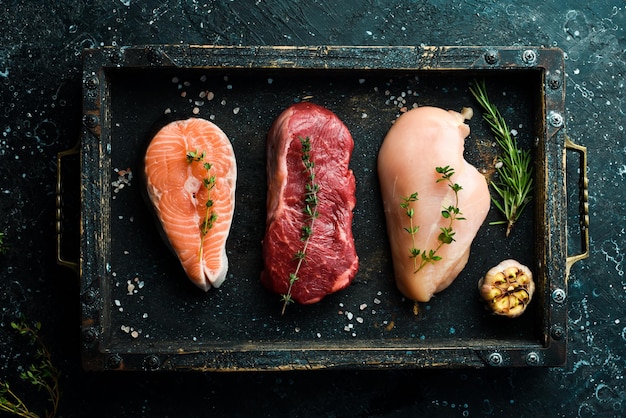 A set of different meats salmon steak beef steak and chicken breast On a black stone background