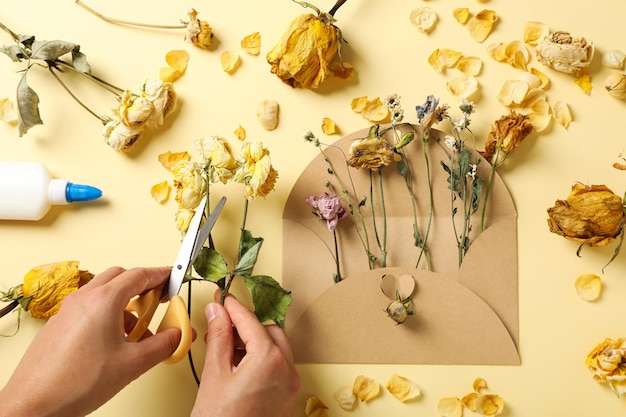 Photo set of dried flower petals envelope glue and scissors in hands on beige background top view