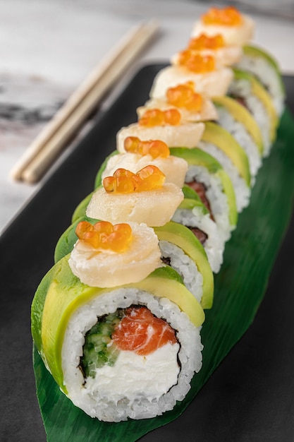 Set of maki rolls with cucumber salmon caviar and green bamboo leaf in a black ceramic plate with chopstick on a bright white textured marble background side view closeup