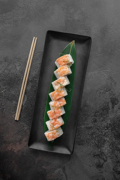 Set of maki rolls with tiger shrimp salmon microgreens and green bamboo leaf in a black ceramic plate with chopstick on a dark gray textured background top view