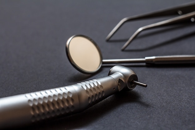 Set of metal dental instruments for dental treatment. High-speed dental handpiece, dental mirror and tweezers on black background. Medical tools. Shallow depth of field. Focus on the bur.