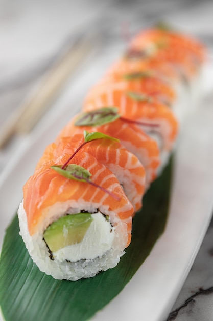 Set of rolls philadelphia classic with microgreens and green bamboo leaf in a white ceramic plate with chopstick on a bright textured marble background side view closeup