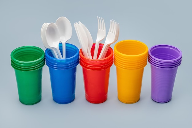 A set, in a row, of multi-colored disposable cups for drinks and cutlery. Grey background.