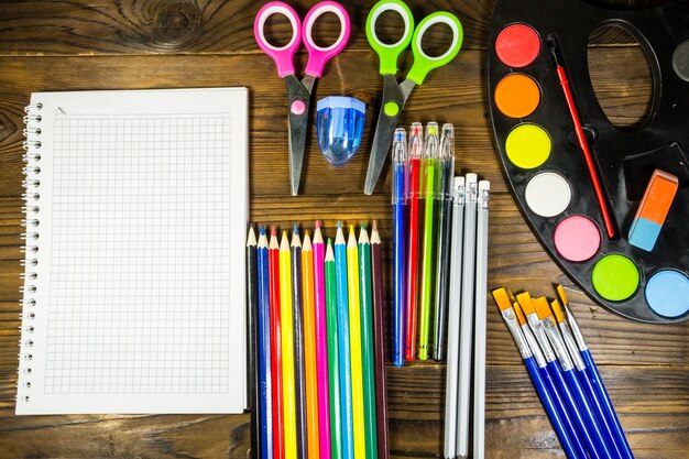 Set of school stationery supplies. Blank notebook, colored pencils, pens, watercolor paints, scissors and other items on wooden desk. Back to school concept