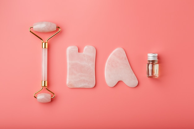 A set of tools for face Massage technique Gua Sha made of natural rose quartz on a pink background. Roller, jade stone and oil in a glass jar for face and body care.