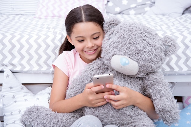 Set up alarm clock or notification Girl child sit on bed with teddy bear in her bedroom Kid prepare to go to bed Girl kid relax hug plush teddy bear toy Child smartphone set up morning alarm