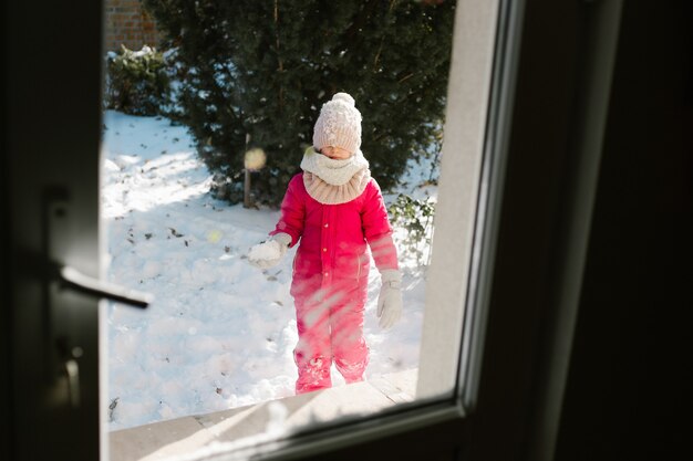 Seven-year-old cute girl in winter clothes is standing outside the door, on the street with snow in her hands and looking into the house smiling.