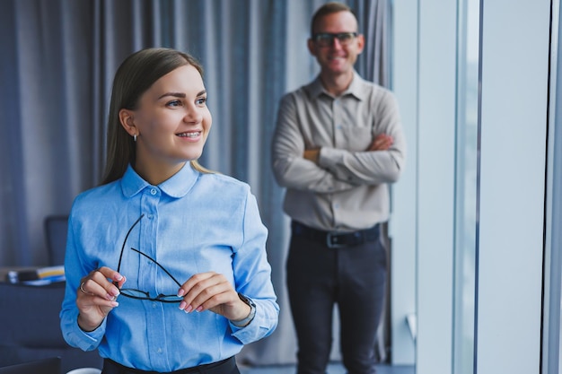 Several friendly colleagues are standing by the office window A business woman in glasses stands on the background of a man in a shirt Selective focus Portrait of business people