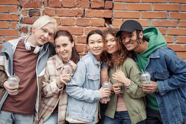 Several happy intercultural teenagers with soda standing against brick wall