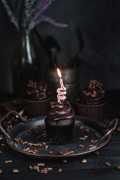 Several muffins or cupcakes with chocolate shaped cream at black table. Festive candle burns on a chocolate cake.