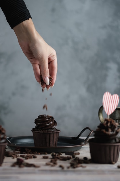 Several muffins or cupcakes with chocolate shaped cream at white table. A card in the form of a heart for St. Valentine's Day. A woman's hand crumbles grated chocolate onto a cake.