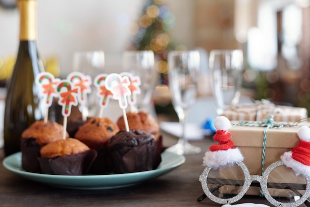 Several tasty homemade muffins with small xmas cards on plate, bottle of champagne, Santa eyeglasses, flutes and giftboxes on table