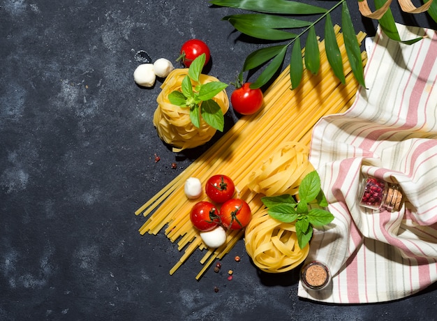 Several types of dry pasta with vegetables and herbs on a dark stone tab