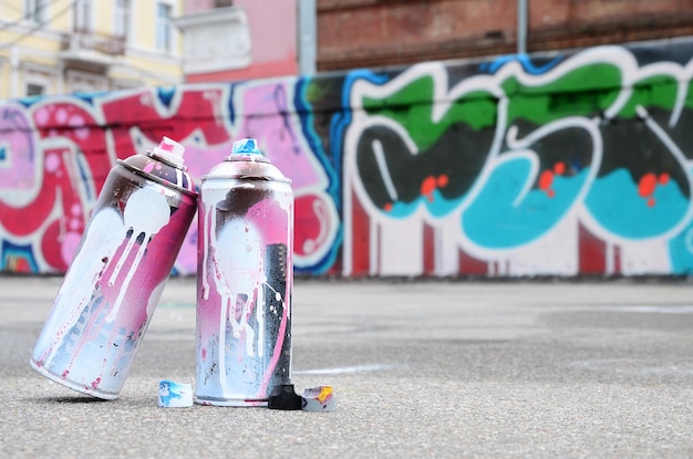 Several used spray cans with pink and white paint and caps for spraying paint under pressure is lies on the asphalt near the painted wall in colored graffiti drawings