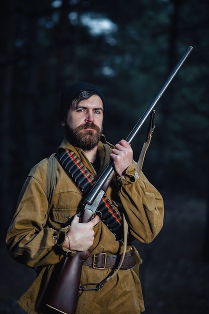 Severe brutal bearded man hunter in a black hat and khaki jacket in a long cloak holds a gun in his hand directed towards the forest