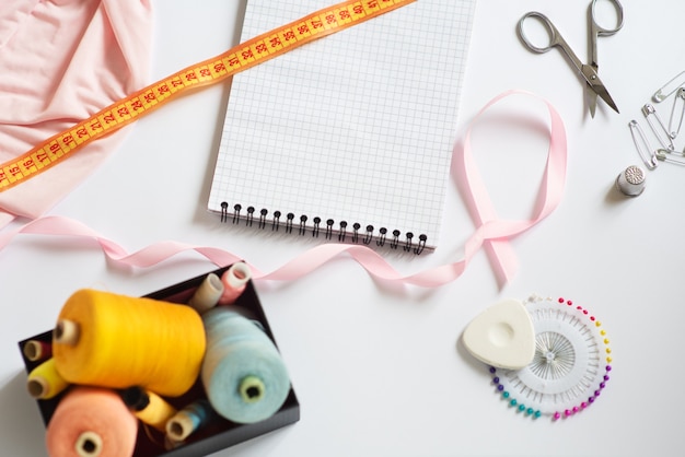 Sewing materials - a box with threads of bright colors, a pink ribbon, cotton, measuring tape on a white background. Frame, space for text